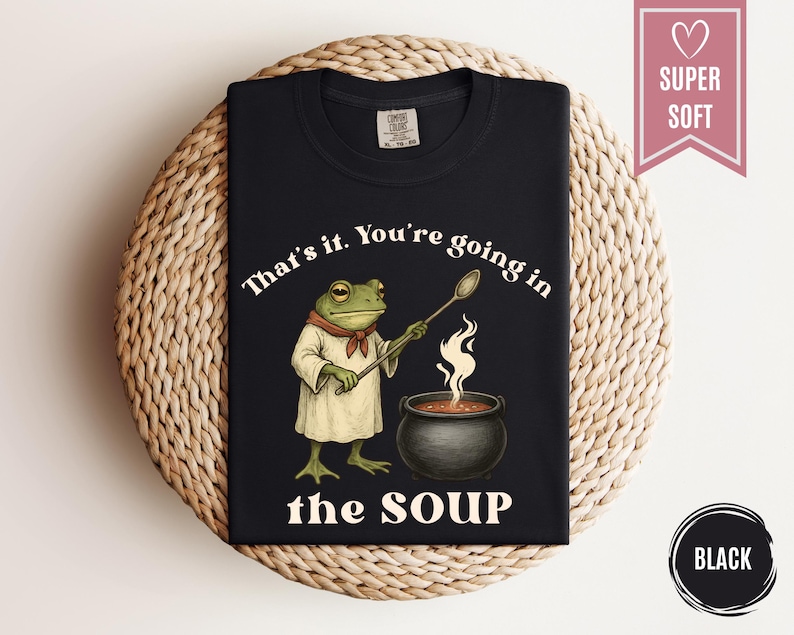 May include: Black t-shirt featuring a cartoon frog chef stirring a pot of soup. The text on the shirt reads "That's it. You're going in the SOUP." A pink banner with a heart and the words "SUPER SOFT" is in the upper right corner.