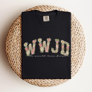 K&ouml;nnte beinhalten: Schwarzes T-Shirt mit den Buchstaben "WWJD" in einem floralen Design, mit rosa und wei&szlig;en Blumen und gr&uuml;nen Bl&auml;ttern. Unter den Buchstaben steht der Text "We would love first". Das Shirt ist auf einer gewebten, kreisf&ouml;rmigen Oberfl&auml;che gefaltet.