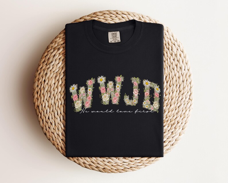 K&ouml;nnte beinhalten: Schwarzes T-Shirt mit den Buchstaben "WWJD" in einem floralen Design, mit rosa und wei&szlig;en Blumen und gr&uuml;nen Bl&auml;ttern. Unter den Buchstaben steht der Text "We would love first". Das Shirt ist auf einer gewebten, kreisf&ouml;rmigen Oberfl&auml;che gefaltet.