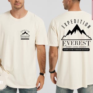 Op de afbeelding: Ivoor kleurig t-shirt met een zwarte afbeelding van een bergketen en het woord "EVEREST". De achterkant van het shirt heeft de tekst "EXPEDITION" boven de bergafbeelding, met "DISNEY'S ANIMAL KINGDOM" en "LEGEND OF THE FORBIDDEN MOUNTAIN" eronder.