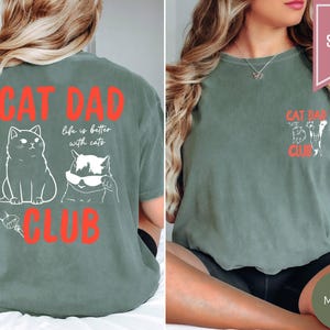 May include: Moss green t-shirt with the text "CAT DAD CLUB" in red. The back of the shirt features two white cat illustrations and the phrase "life is better with cats." The front has a smaller design with cat illustrations and the same text.