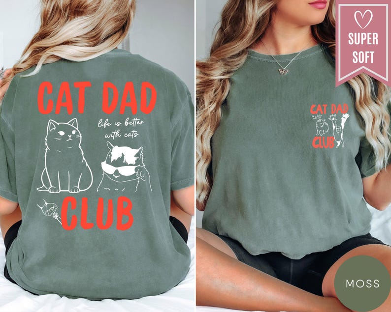 May include: Moss green t-shirt with the text "CAT DAD CLUB" in red. The back of the shirt features two white cat illustrations and the phrase "life is better with cats." The front has a smaller design with cat illustrations and the same text.