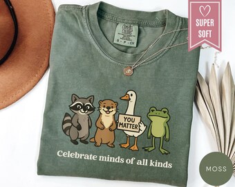 Könnte beinhalten: Moosgrünes T-Shirt mit einer Grafik eines Waschbären, Otters, einer Ente und eines Frosches. Die Ente hält ein Schild mit der Aufschrift "You Matter". Der Text "Celebrate minds of all kinds" ist darunter gedruckt. Eine goldene Halskette befindet sich auf dem Shirt.