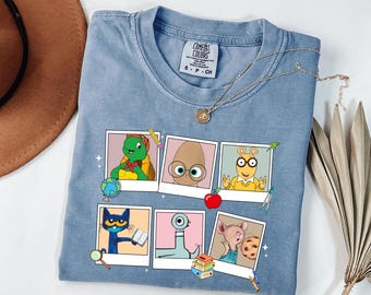 May include: A light blue Comfort Colors t-shirt featuring six cartoon character portraits in polaroid frames. The shirt has a crew neck and short sleeves. A gold necklace with a pendant is draped on the shirt.