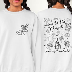 K&ouml;nnte beinhalten: Wei&szlig;es Sweatshirt mit schwarzer Strichzeichnung. Auf der Vorderseite befindet sich ein herzf&ouml;rmiges Medaillon mit der Aufschrift "Mrs. Wilson". Auf der R&uuml;ckseite steht der Satz "going to the Chapel, gonna get married" mit Illustrationen zum Thema Hochzeit.