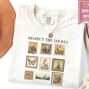 May include: Ivory-colored t-shirt with the text "Respect the Locals" above a series of vintage-style postage stamp illustrations of wildlife, including a bear, eagle, and fox. The shirt is labeled "Super Soft" and is paired with a gold necklace.