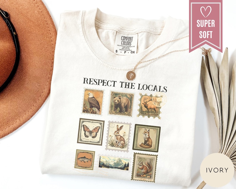 May include: Ivory-colored t-shirt with the text "Respect the Locals" above a series of vintage-style postage stamp illustrations of wildlife, including a bear, eagle, and fox. The shirt is labeled "Super Soft" and is paired with a gold necklace.