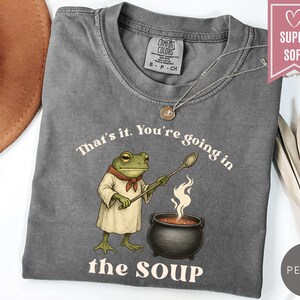 May include: A gray t-shirt with the text "That's it. You're going in the SOUP" and a cartoon frog holding a spoon over a pot of soup. The shirt has a crew neck and short sleeves.