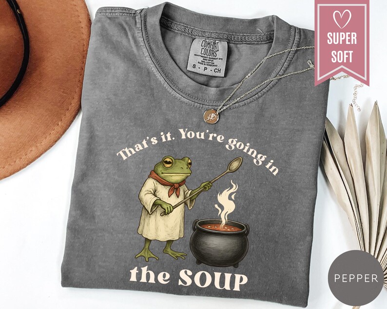 May include: A gray t-shirt with the text "That's it. You're going in the SOUP" and a cartoon frog holding a spoon over a pot of soup. The shirt has a crew neck and short sleeves.