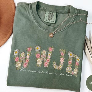 K&ouml;nnte beinhalten: Moosgr&uuml;nes T-Shirt mit floralem "WWJD"-Design und dem Text "We would love first." Das Shirt hat einen Rundhalsausschnitt und kurze &Auml;rmel. Eine goldene Halskette mit Anh&auml;nger liegt &uuml;ber dem Shirt.