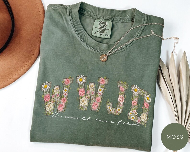 K&ouml;nnte beinhalten: Moosgr&uuml;nes T-Shirt mit floralem "WWJD"-Design und dem Text "We would love first." Das Shirt hat einen Rundhalsausschnitt und kurze &Auml;rmel. Eine goldene Halskette mit Anh&auml;nger liegt &uuml;ber dem Shirt.