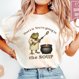 May include: Ivory-colored t-shirt featuring a frog chef with a spoon, standing next to a bubbling pot of soup. The text "That's it. You're going in the SOUP" is printed above and below the frog illustration. A "SUPER SOFT" banner is in the upper right.