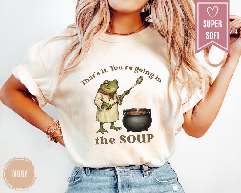 May include: Ivory-colored t-shirt featuring a frog chef with a spoon, standing next to a bubbling pot of soup. The text "That's it. You're going in the SOUP" is printed above and below the frog illustration. A "SUPER SOFT" banner is in the upper right.