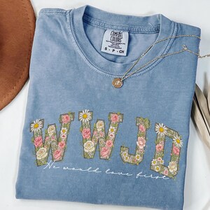 K&ouml;nnte beinhalten: Hellblaues T-Shirt mit den Buchstaben "WWJD", verziert mit rosa, wei&szlig;en und gr&uuml;nen Blumenmustern. Darunter steht der Text "We would love first". Eine goldene Halskette und ein brauner Hut sind ebenfalls zu sehen.