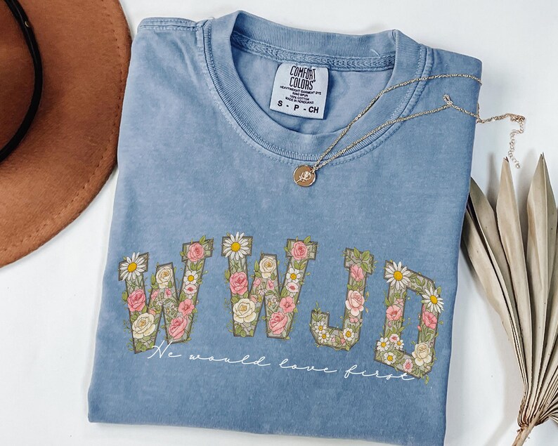 K&ouml;nnte beinhalten: Hellblaues T-Shirt mit den Buchstaben "WWJD", verziert mit rosa, wei&szlig;en und gr&uuml;nen Blumenmustern. Darunter steht der Text "We would love first". Eine goldene Halskette und ein brauner Hut sind ebenfalls zu sehen.