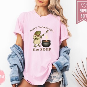 May include: A light pink t-shirt with the text "That's it. You're going in the SOUP" and a cartoon frog chef stirring a pot of soup. The shirt is made of a soft material.