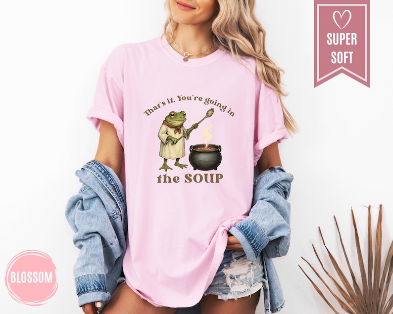 May include: A light pink t-shirt with the text "That's it. You're going in the SOUP" and a cartoon frog chef stirring a pot of soup. The shirt is made of a soft material.
