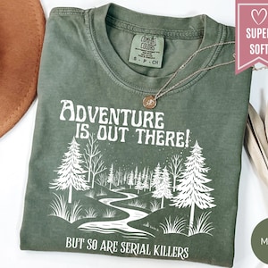May include: Moss green t-shirt with the text "Adventure is Out There! But So Are Serial Killers" in white. The graphic features a forest scene with a winding river. A gold necklace is on the shirt.