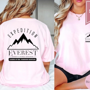 Op de afbeelding: Een lichtroze t-shirt met de tekst "EXPEDITION EVEREST" en een bergafbeelding. Het shirt heeft ook de tekst "DISNEY'S ANIMAL KINGDOM LEGEND OF THE FORBIDDEN MOUNTAIN". Het shirt is gelabeld met "SUPER SOFT" en "BLOSSOM".