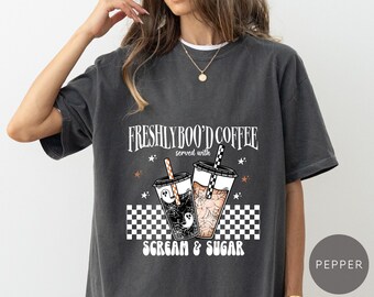 May include: Dark gray t-shirt with a graphic that reads "FRESHLY BOO'D COFFEE served with SCREAM & SUGAR." The design features two coffee cups with Halloween-themed illustrations, including ghosts and spiderwebs. The shirt has a relaxed fit.