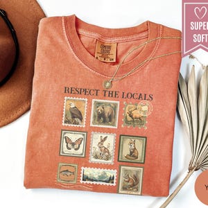 May include: A yam-colored t-shirt with the text "Respect the Locals" and images of wildlife stamps. The shirt has a crew neck and short sleeves. A gold necklace is draped across the shirt. The shirt is labeled "Super Soft".