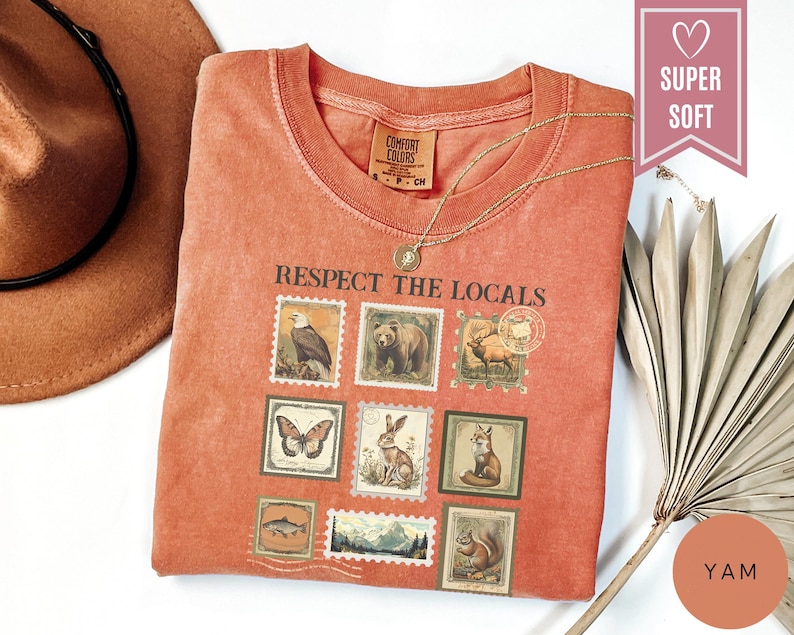 May include: A yam-colored t-shirt with the text "Respect the Locals" and images of wildlife stamps. The shirt has a crew neck and short sleeves. A gold necklace is draped across the shirt. The shirt is labeled "Super Soft".