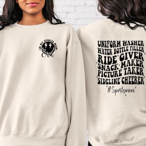 Pode incluir: Uma camisa de moletom cor creme com texto preto que diz "In my sports mom era" na frente e "Uniform washer, water bottle filler, ride giver, snack maker, picture taker, sideline cheerleader #Sportsmom" nas costas.