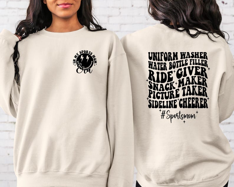 Pode incluir: Uma camisa de moletom cor creme com texto preto que diz "In my sports mom era" na frente e "Uniform washer, water bottle filler, ride giver, snack maker, picture taker, sideline cheerleader #Sportsmom" nas costas.