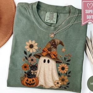 May include: A moss green t-shirt featuring a graphic of a white ghost wearing a brown patchwork witch hat, sitting next to a black cat on a pumpkin, surrounded by orange and brown flowers. The shirt has a Comfort Colors tag and a necklace.