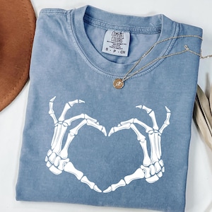 May include: A blue Comfort Colors t-shirt with a white skeleton hand heart design. A gold necklace with a pendant is draped across the shirt. The shirt is folded and has a tag that reads "Comfort Colors".