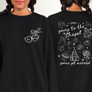 K&ouml;nnte beinhalten: Schwarzes Sweatshirt mit wei&szlig;er Strichzeichnung. Auf der Vorderseite befinden sich zwei verbundene herzf&ouml;rmige Designs mit dem Text "Mrs. Wilson". Auf der R&uuml;ckseite steht der Satz "going to the Chapel, gonna get married" mit Illustrationen zum Thema Hochzeit.