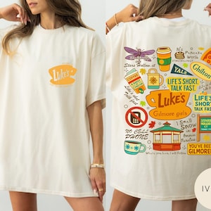 May include: Ivory-colored t-shirt featuring the text "Luke's" on the front and a colorful graphic on the back with the text "Gilmore Girls", coffee cups, and other themed elements. The shirt has a relaxed fit.