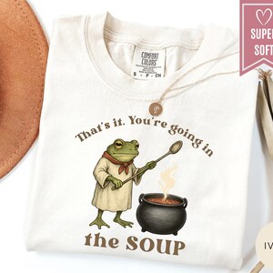May include: Ivory-colored t-shirt with the text "That's it. You're going in the SOUP." A cartoon frog wearing a chef's outfit stirs a pot of soup. The shirt has a "SUPER SOFT" label.