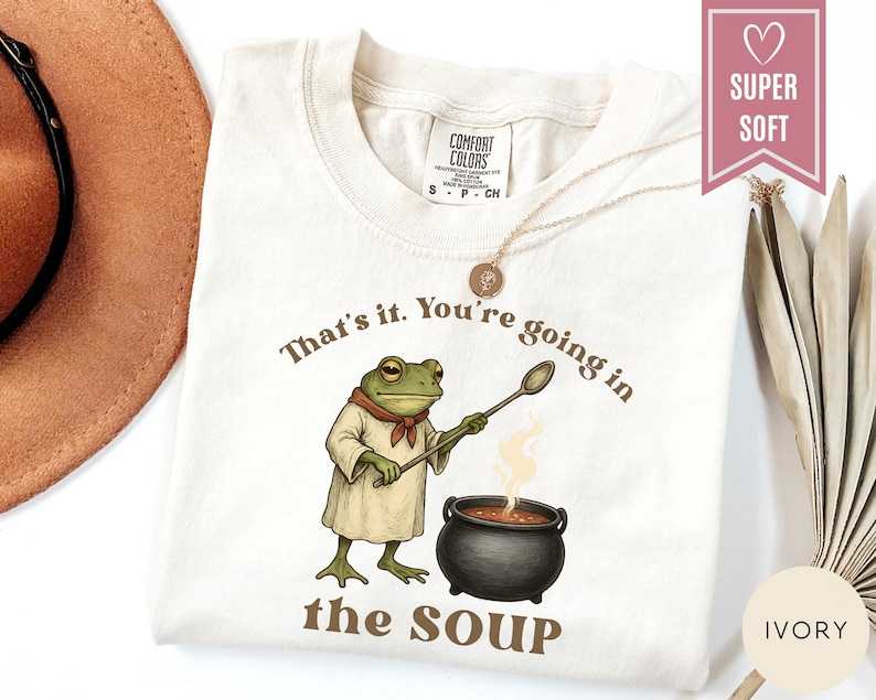 May include: Ivory-colored t-shirt with the text "That's it. You're going in the SOUP." A cartoon frog wearing a chef's outfit stirs a pot of soup. The shirt has a "SUPER SOFT" label.