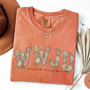 K&ouml;nnte beinhalten: Korallfarbenes T-Shirt mit den Buchstaben "WWJD", die mit floralen Designs gef&uuml;llt sind. Der Text "We would love first" steht unter den Buchstaben. Eine goldene Halskette und ein brauner Hut liegen neben dem Shirt.