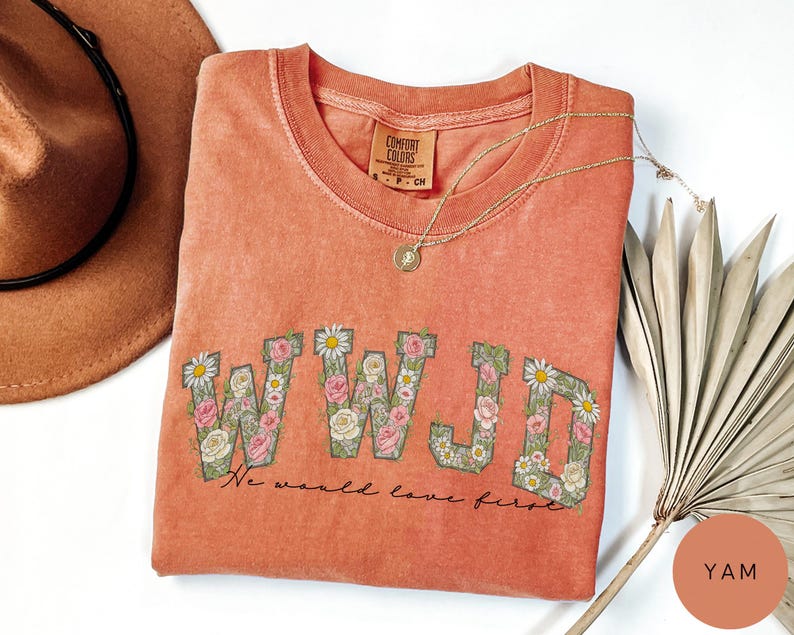 K&ouml;nnte beinhalten: Korallfarbenes T-Shirt mit den Buchstaben "WWJD", die mit floralen Designs gef&uuml;llt sind. Der Text "We would love first" steht unter den Buchstaben. Eine goldene Halskette und ein brauner Hut liegen neben dem Shirt.