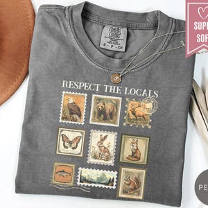 May include: A gray t-shirt with the phrase "Respect the Locals" and postage stamp-style illustrations of wildlife. The shirt has a crew neck and short sleeves. A silver necklace with a pendant is on the shirt.