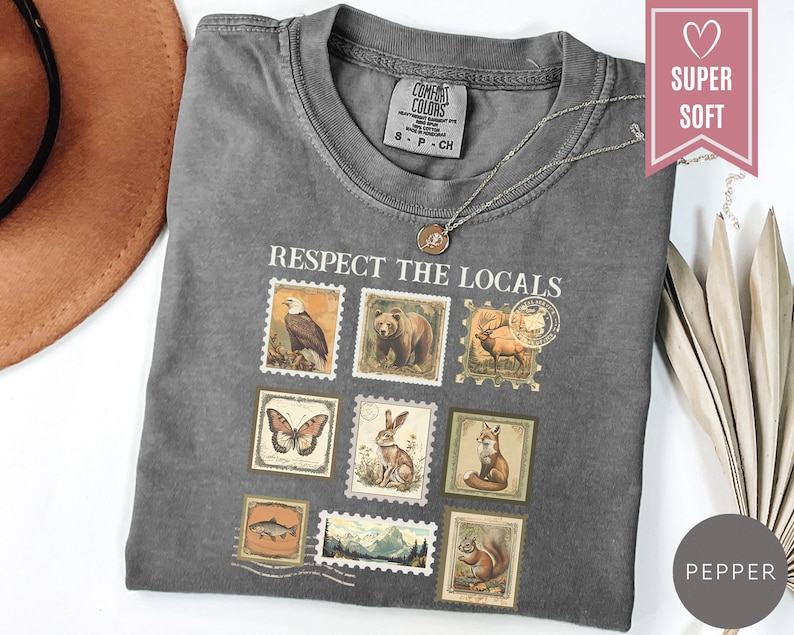 May include: A gray t-shirt with the phrase "Respect the Locals" and postage stamp-style illustrations of wildlife. The shirt has a crew neck and short sleeves. A silver necklace with a pendant is on the shirt.