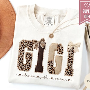 May include: Ivory-colored t-shirt with the word "GIGI" in a leopard print design, accented with brown patches and bows. The names "olivia", "josh", and "emma" are printed below. The shirt is labeled "SUPER SOFT".