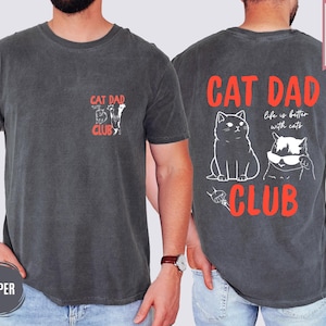 May include: Pepper-colored t-shirt with "CAT DAD CLUB" text and cat illustrations. The back features a larger design with two cats and the phrase "life is better with cats". The shirt is labeled "SUPER SOFT".