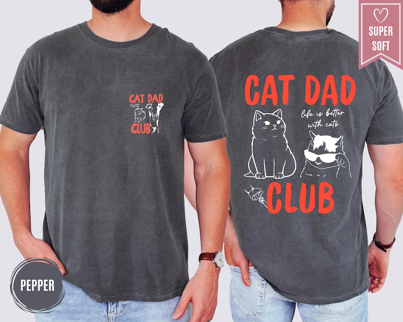 May include: Pepper-colored t-shirt with "CAT DAD CLUB" text and cat illustrations. The back features a larger design with two cats and the phrase "life is better with cats". The shirt is labeled "SUPER SOFT".