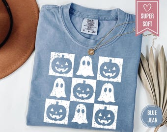 May include: Blue jean colored t-shirt with a white Halloween-themed design. The design features a grid of jack-o'-lanterns and ghosts. The shirt has a crew neck and short sleeves. A gold necklace is draped across the shirt.