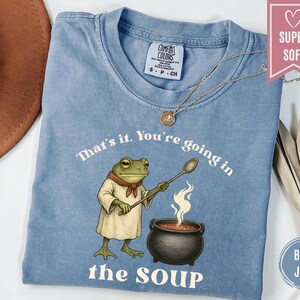 May include: Blue jean Comfort Colors t-shirt with a frog chef graphic holding a spoon over a pot of soup. The text reads "That's it. You're going in the soup."