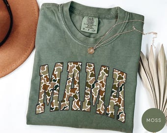 May include: Moss green t-shirt with the word "MAMA" in a camouflage pattern. The shirt has a crew neck and short sleeves. A gold necklace rests on the shirt. A brown hat and dried leaves are in the background.