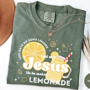 Puede incluir: Camiseta verde musgo con el texto "Life gave me some lemons but my Jesus He be makin' lemonade". El gr&aacute;fico incluye una gran rodaja de lim&oacute;n y una bebida de limonada. Un collar dorado est&aacute; drapeado sobre la camiseta.