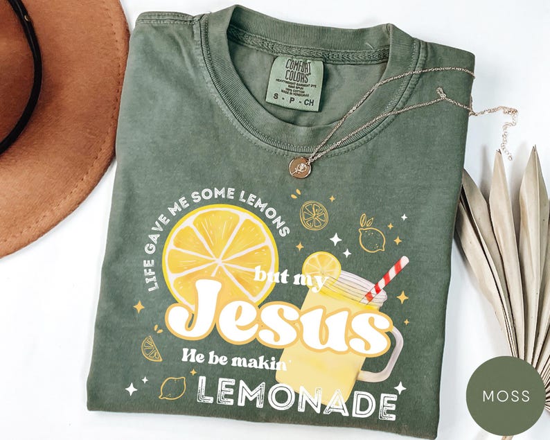 Puede incluir: Camiseta verde musgo con el texto "Life gave me some lemons but my Jesus He be makin' lemonade". El gr&aacute;fico incluye una gran rodaja de lim&oacute;n y una bebida de limonada. Un collar dorado est&aacute; drapeado sobre la camiseta.