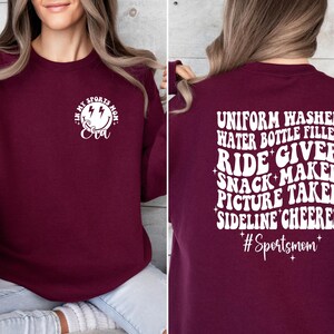 Pode incluir: Camisola de cor bord&ocirc; com texto branco que diz "Eu sou a m&atilde;e esportiva da Eva" na frente e "Lavadora de uniformes, enchimento de garrafas de &aacute;gua, acompanhante, fabricante de lanches, fot&oacute;grafa, animadora de torcida #M&atilde;eEsportiva" nas costas.