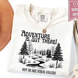 May include: Ivory-colored t-shirt with the text "Adventure is Out There!" above a black and white illustration of a forest scene. The text "But So Are Serial Killers" is below the illustration. The shirt has a crew neck and short sleeves.
