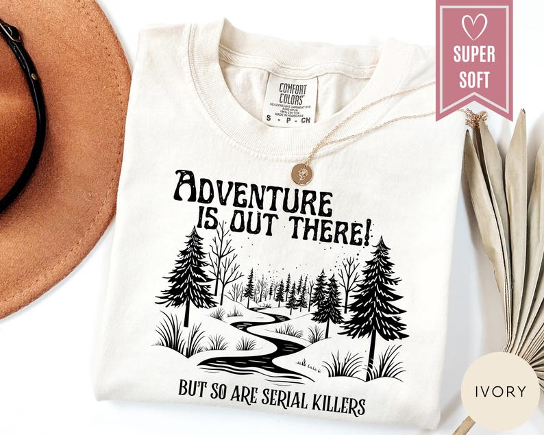 May include: Ivory-colored t-shirt with the text "Adventure is Out There!" above a black and white illustration of a forest scene. The text "But So Are Serial Killers" is below the illustration. The shirt has a crew neck and short sleeves.