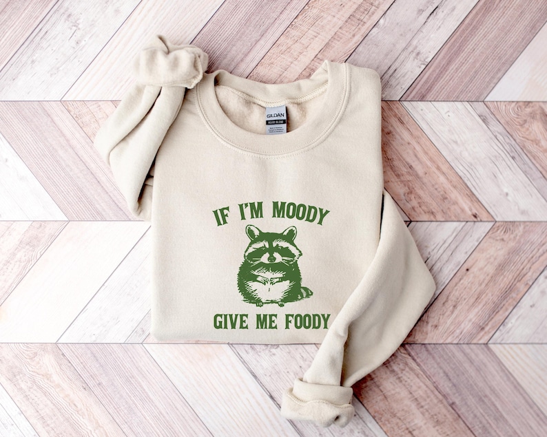 May include: A cream-coloured sweatshirt with the text "If I'm Moody Give Me Foody" printed in green. A cartoon raccoon with a green bandana is pictured above the text.